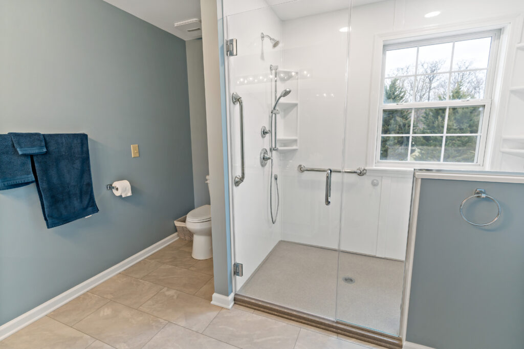 A newly renovated modern bathroom with a frameless glass shower enclosure, blue-gray walls, and neutral tile flooring, photographed during daylight hours.