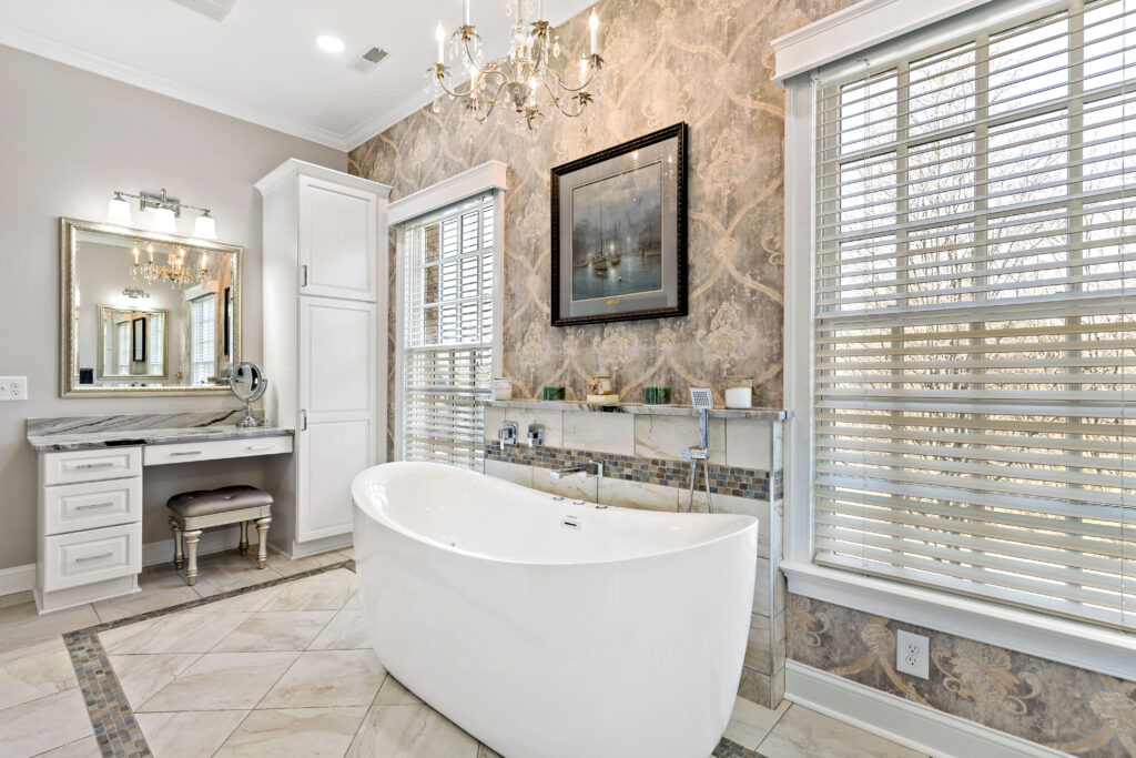 A luxurious modern bathroom featuring a freestanding white bathtub, dual vanities with mirrors, a crystal chandelier, and elegant tile accents with shuttered windows.