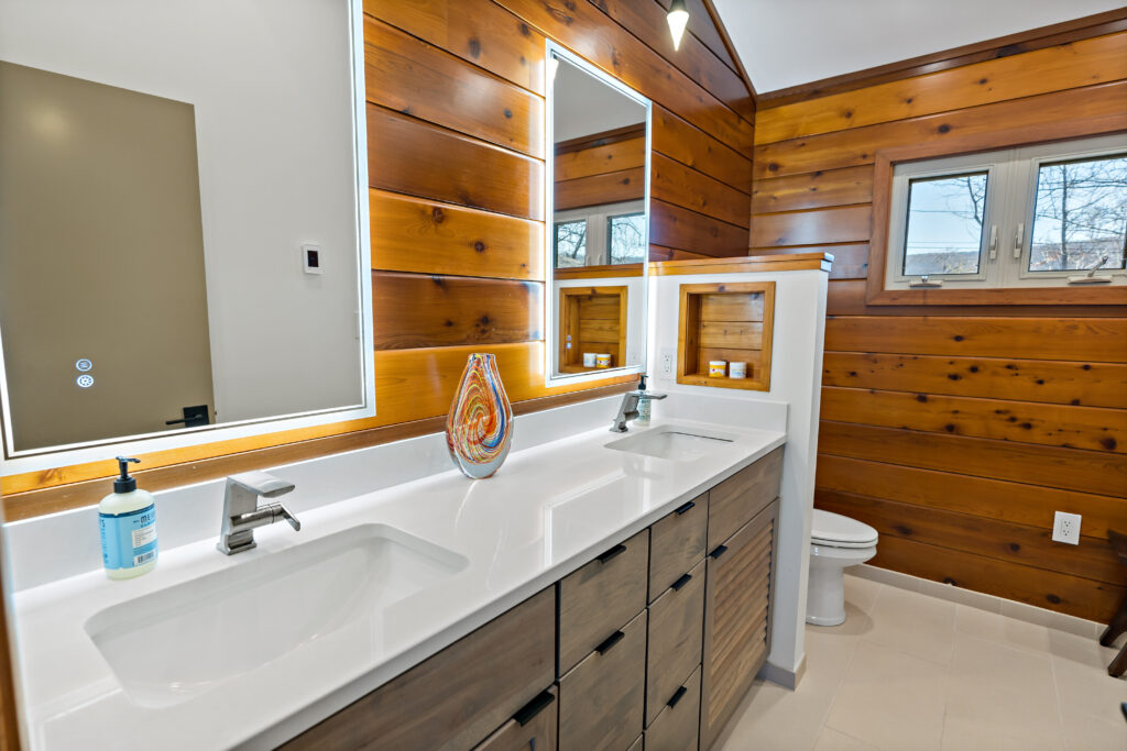 A modern bathroom features a double-sink vanity with dark cabinetry and a white countertop, complemented by warm wooden shelving and paneling throughout the space.