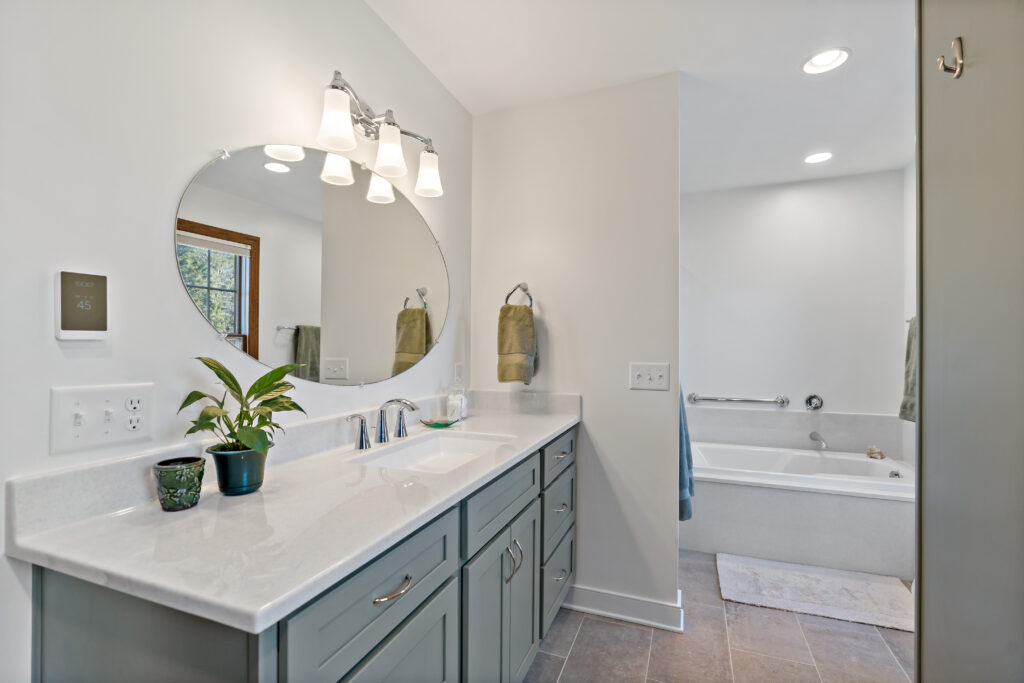 A modern, bright bathroom features a gray vanity with white countertop, large mirror with lighting, and an adjacent soaking tub in a clean, minimalist design.
