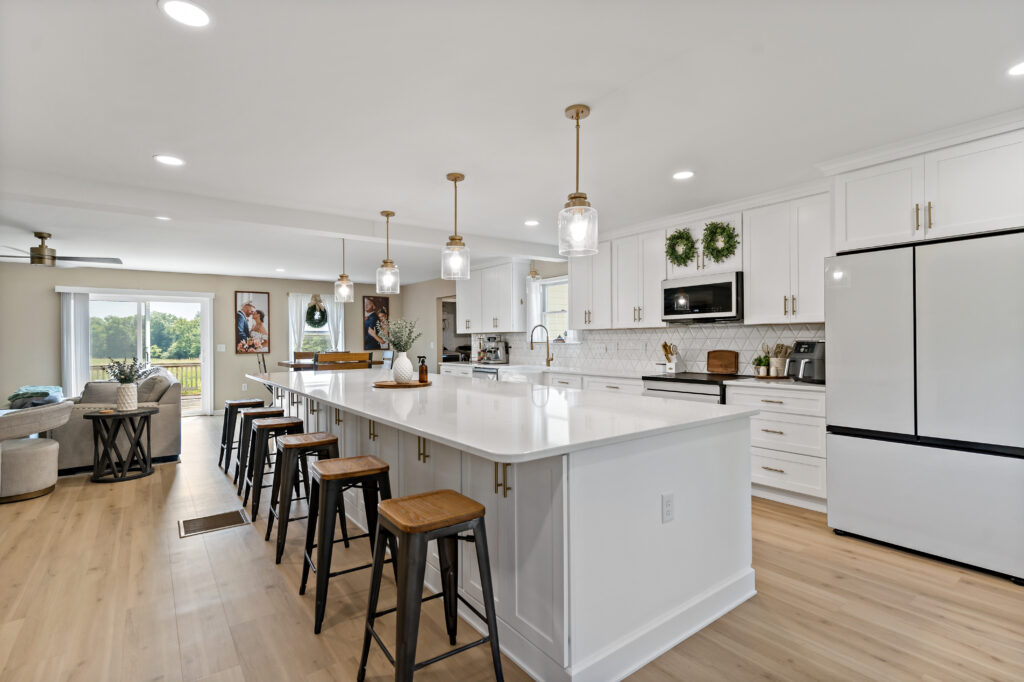 A bright, modern kitchen featuring a large white island with bar seating, stainless steel appliances, white cabinetry, pendant lighting, and an open floor plan connecting to a living area.