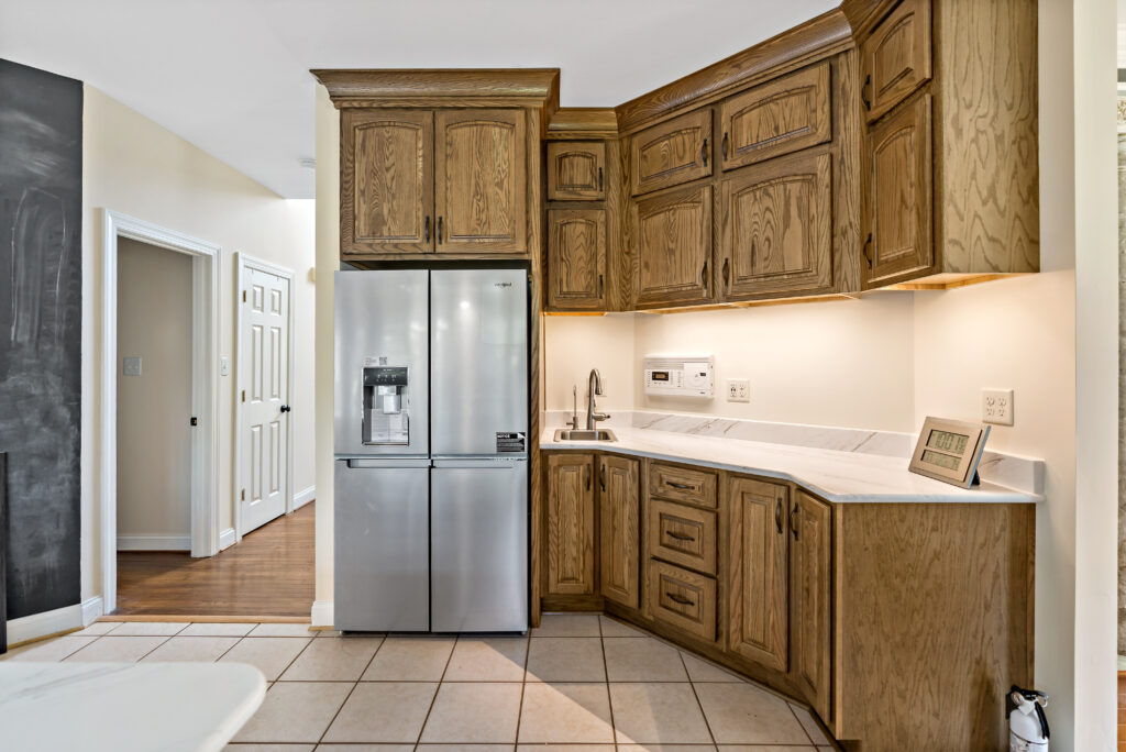 A modern kitchen interior featuring warm wooden cabinetry, stainless steel appliances, and neutral tile flooring with white countertops.