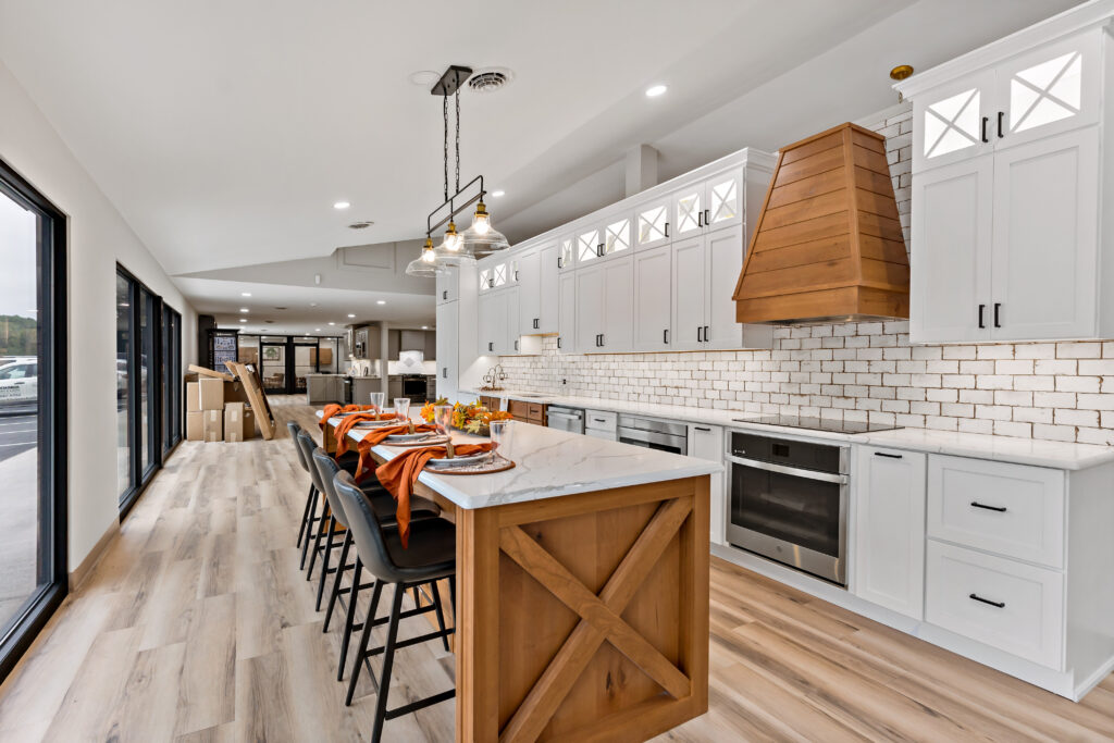 A modern, open-concept kitchen featuring white cabinetry, a wooden island with bar seating, pendant lighting, and natural light from large windows.