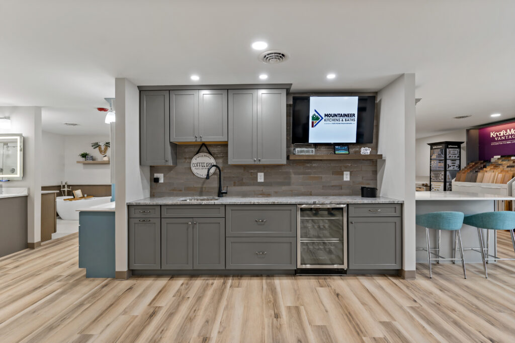 A modern kitchen with gray cabinetry, wooden flooring, and a mounted television above the counter area features an open floor plan with bar seating.