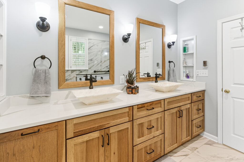 A modern bathroom vanity features warm wood cabinetry with dual mirrors, white countertops, and contemporary fixtures in a bright, clean space.
