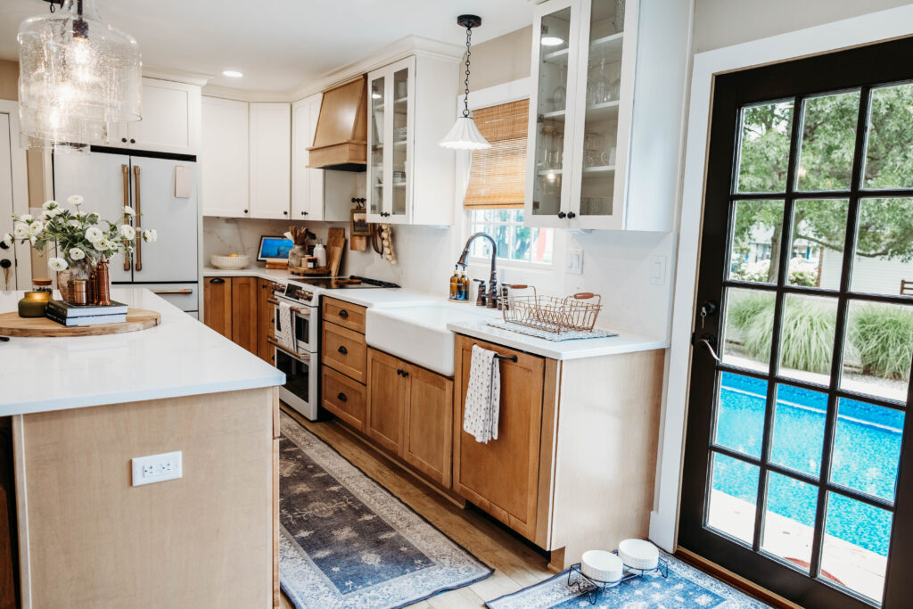 A bright, contemporary kitchen features white cabinetry with wood accents, a large island, white countertops, and expansive glass doors overlooking a pool.