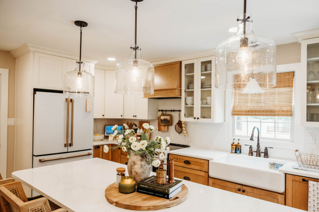 A bright, contemporary kitchen features white cabinetry, warm wood accents, pendant lighting, and a marble-topped island with fresh floral decoration.