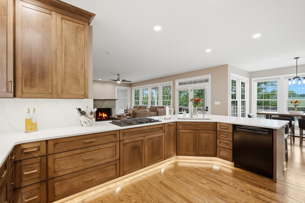A spacious, contemporary kitchen features rich wooden cabinetry, white countertops, a large island, and abundant natural light from multiple windows overlooking landscaped grounds.