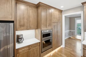A contemporary kitchen features warm wood cabinetry, stainless steel appliances, white subway tile backsplash, and hardwood flooring with an open doorway to adjacent rooms.