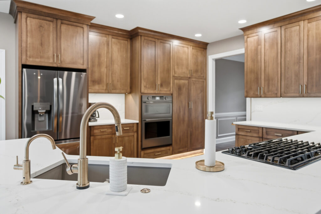 A luxurious modern kitchen features warm wood cabinetry, white marble countertops, stainless steel appliances, and brass fixtures in a bright, professionally designed space.