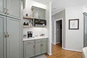 A modern kitchen with sage green cabinetry, open shelving, brass hardware, and white countertops in a bright residential interior.