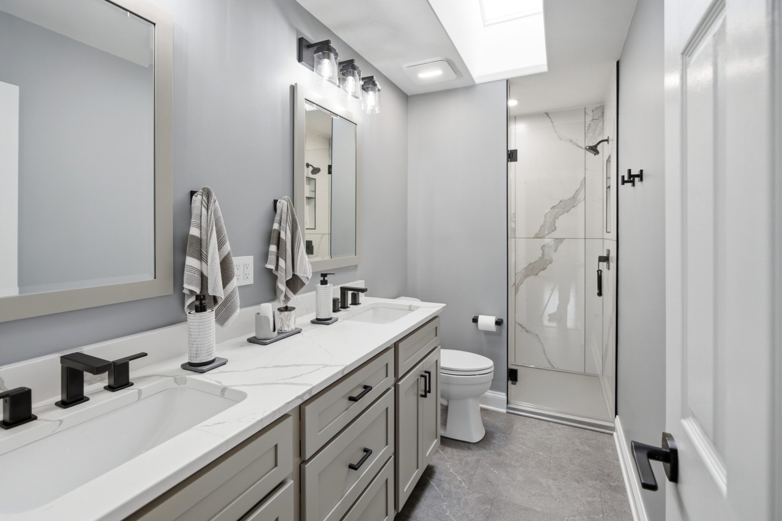 A modern gray bathroom features a double-sink vanity with marble countertop, framed mirrors, black fixtures, and a glass-enclosed shower with marble tile.
